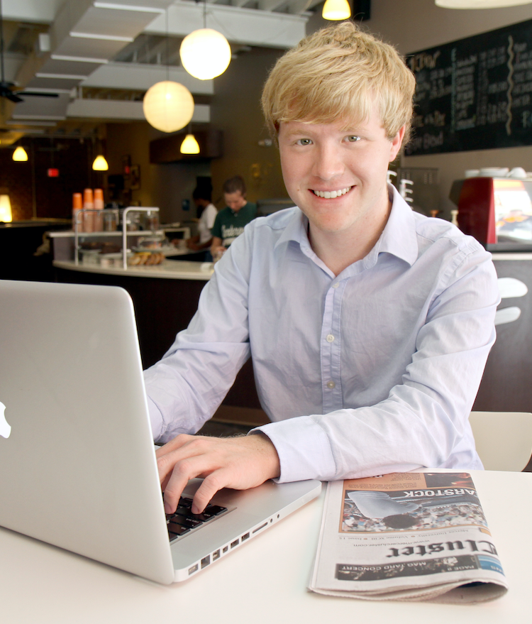 Student using a laptop in a coffee shop with a Mercer Cluster newspaper on the table